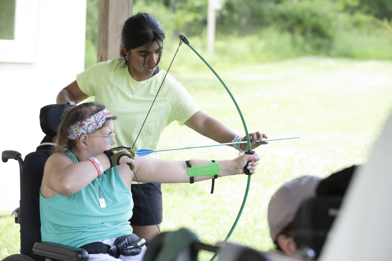Camp Cranium participant Angelica Zander aims a bow and arrow while sitting in a wheelchair, as camp counselor Anvitha Tharra stands next to her and helps her learn to use the bow and arrow.
