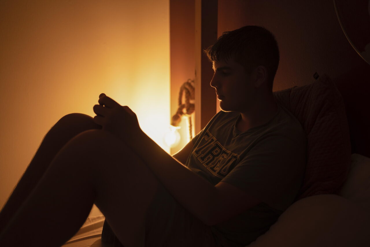 A teen boy is shown sitting up in his bed, looking at his phone. The room is dark except for dim lamplight.