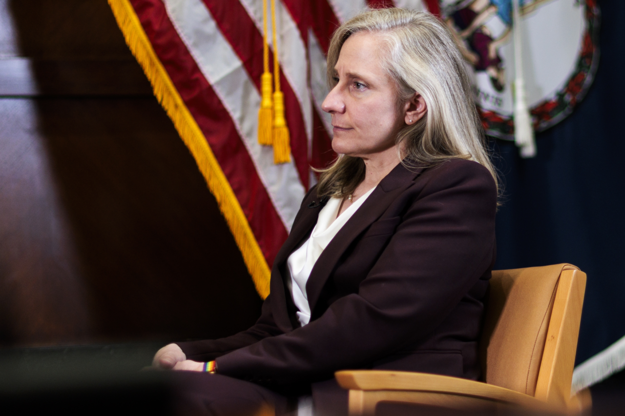 A blonde woman in a dark suit sits in a chair and listens. The American flag and the Virginia state flag are visible in the background.