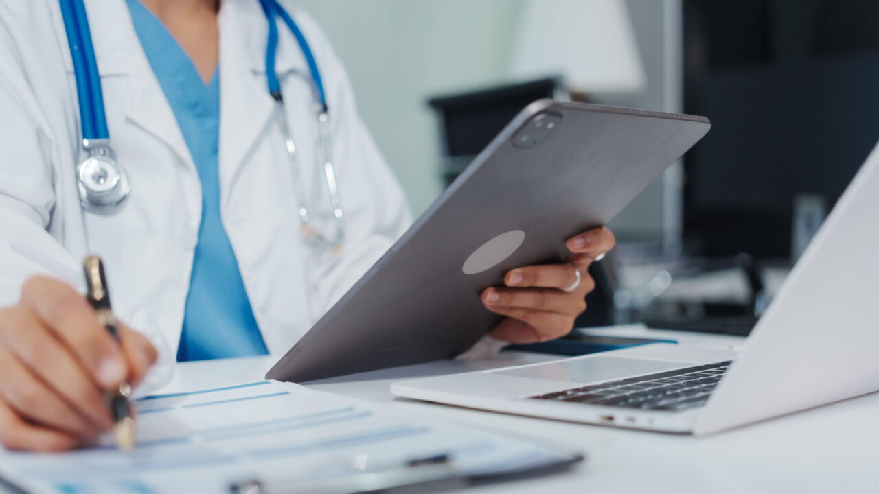 A light-skinned medical professional positions behind a table holds a tablet in one hand and a pen to a blurred medical chart in the other. They're wearing blur scrubs with a white medical coat and a stethoscope around their neck. The photo cuts off above the neck.