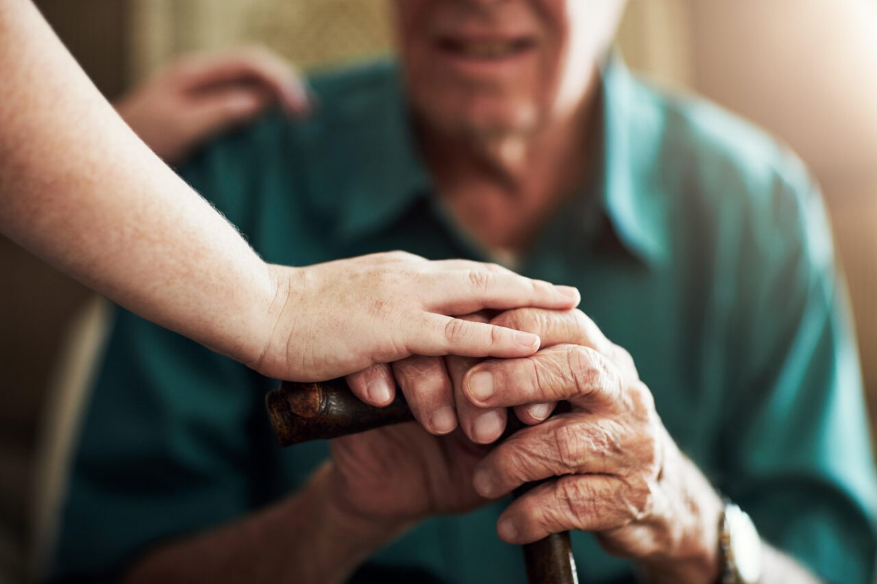 A caregiver holds the hand of an older adult. Staffing shortages are among the challenges facing nursing homes as facilities work to meet residents’ daily care needs.