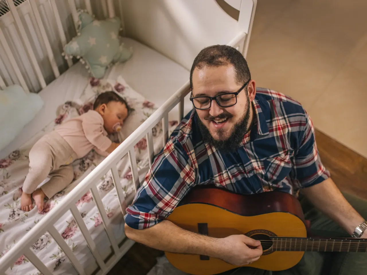 A young father lulls his baby to sleep while playing the guitar and singing.