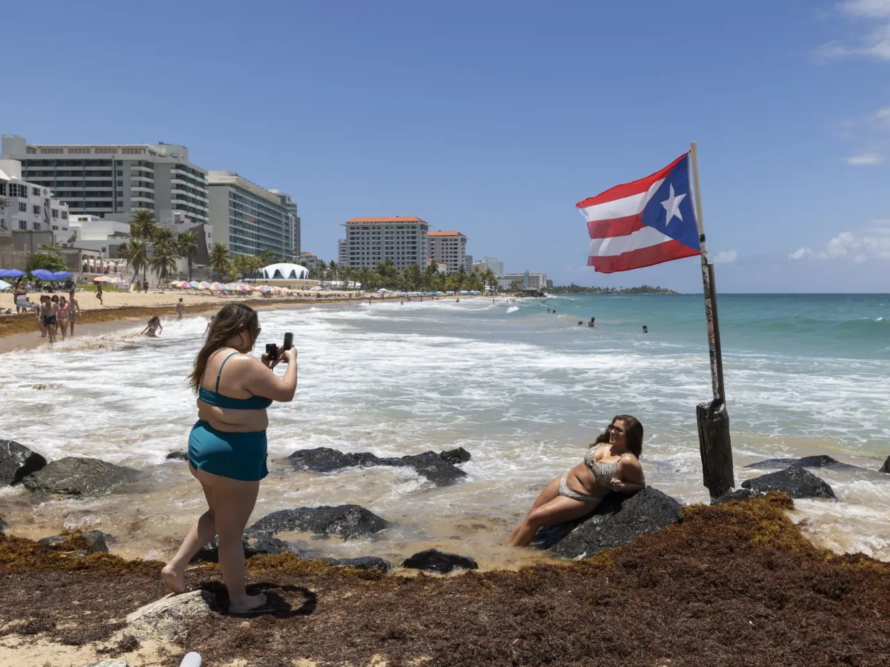 A woman poses for photos next to a Puerto Rican flag along the beach in Condado, Puerto Rico, as Hurricane Erin approaches, Friday, Aug. 15, 2025.