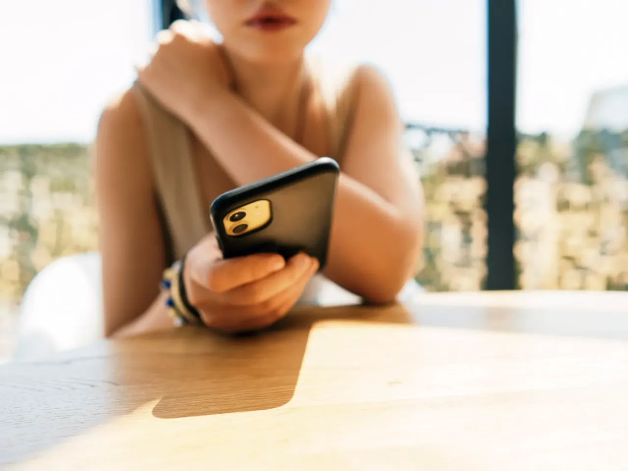 A young woman sits at a sunlit table, holding a smartphone with a soft focus background. The scene conveys casual connectivity, focus on the device, and a relaxed moment of everyday life.