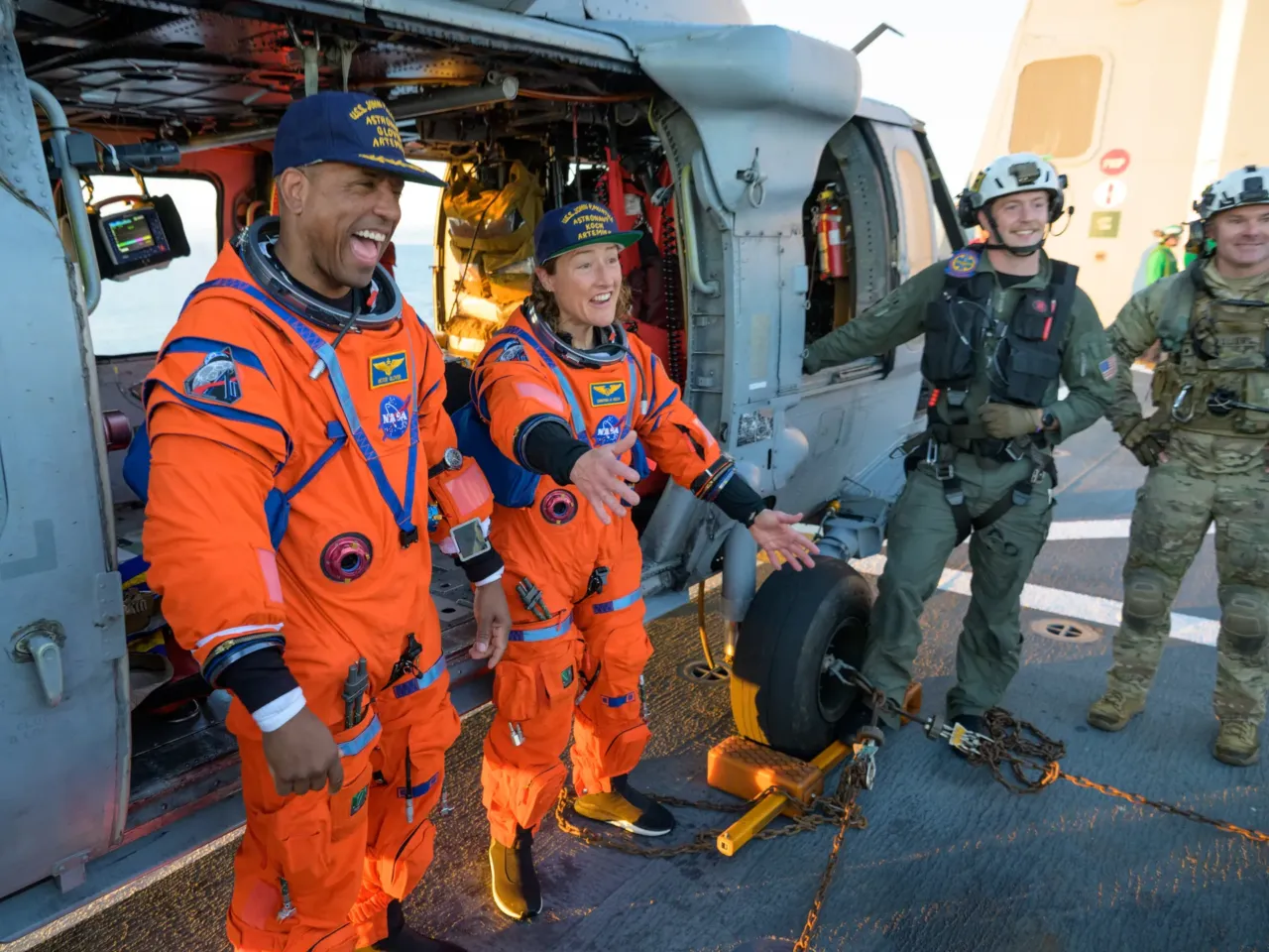NASA astronaut Victor Glover, Artemis II pilot, left, and NASA astronaut Christina Koch, Artemis II mission specialist are seen sitting on a Navy MH-60 Seahawk from Helicopter Sea Combat Squadron (HSC) 23 on the flight deck of USS John P. Murtha.